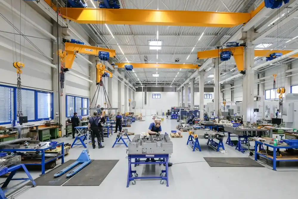 Bright, modern production hall for toolmaking and special machine engineering featuring yellow overhead cranes, workstations, and technicians working on assemblies.