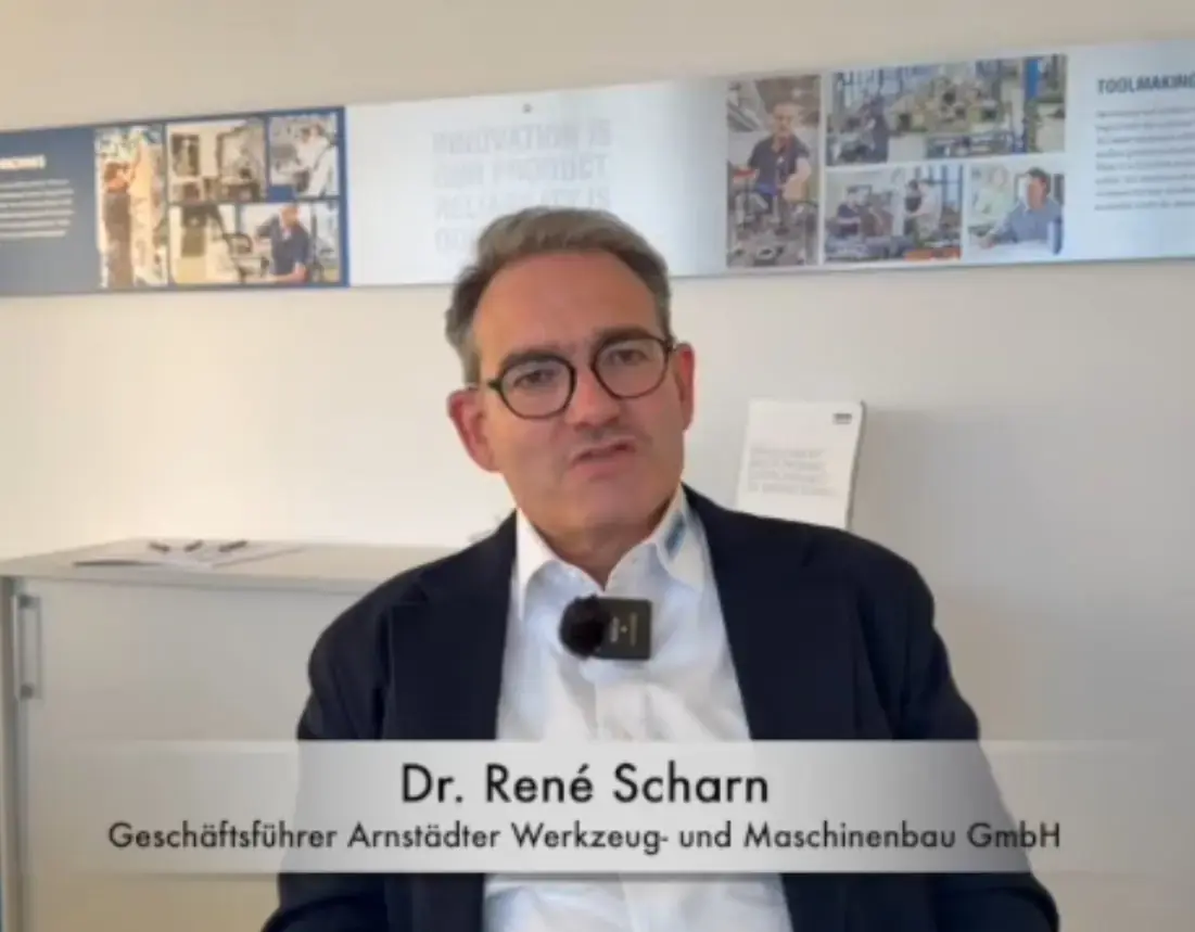 Portrait of Dr. René Scharn in an interview for an AWM corporate video, set against a photo wall displaying company slogans and manufacturing imagery.